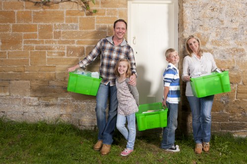 A family of four standing outside a stone and brick building, each holding a bright green plastic container used for rubbish or waste collection. The father, dressed in a checkered shirt and jeans, stands on the left smiling broadly. Next to him is a young girl with long hair, wearing a grey sweater and jeans, holding a container at waist level. To the right, a boy in a striped blue and white shirt and jeans holds a container, standing beside a woman dressed in a light-colored blouse and jeans, who also holds a green waste bin. They are positioned on a small grassy area next to a paved pathway, with the building's wall serving as the background. The wall features a combination of stonework and brickwork, indicating a typical residential or historic structure. The scene appears to be captured on a bright day, with natural lighting illuminating the group and the surroundings, aligning with services provided by Belsize Park House Clearance for rubbish removal and house clearance within the local area near London, England.