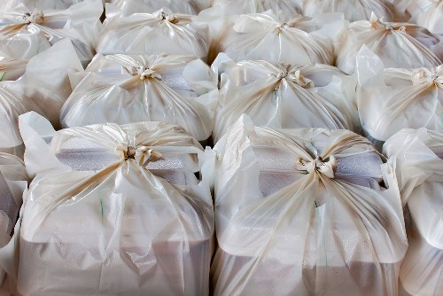 A group of tightly packed white plastic bags filled with household rubbish, tied securely at the top with beige twist ties, arranged on a flat surface in an outdoor or warehouse setting. The bags are made of thin, semi-transparent plastic, allowing faint glimpses of their contents, which appear to be mixed waste materials. The surface beneath the bags is not visible, but the background suggests an industrial environment appropriate for rubbish collection or house clearance services. The image highlights the typical packaging used during rubbish removal processes in Belsize Park or surrounding locations, emphasizing the importance of proper waste containment before disposal or recycling. The bags are orderly lined up, predominantly occupying the central and foreground areas of the image, conveying a sense of readiness for collection by a professional rubbish removal company like Belsize Park House Clearance.