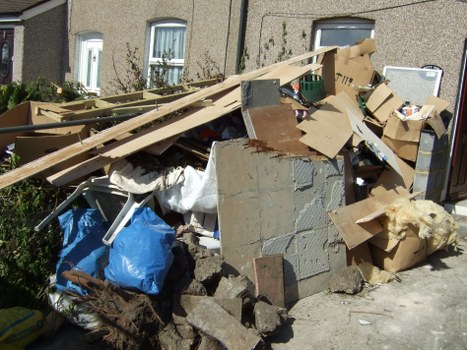 A large pile of mixed waste materials is situated on a paved driveway in an outdoor area, adjacent to residential buildings. The heap contains various types of rubbish, including broken wooden planks, corrugated cardboard boxes, plastic bags, and fragments of concrete or brick. The cardboard packaging appears crushed and torn, with some boxes partially obscured beneath other debris. A white tarp is draped over part of the rubbish to the left, and a green garden spade is visible among the materials. The backdrop features a row of attached houses with light-colored exterior walls, blue window frames, and small front gardens with bushes. The scene suggests a typical residential rubbish clearance, possibly carried out by Belsize Park House Clearance, to remove household waste from a property in the local area near Belsize Park. The natural daylight highlights the cluttered arrangement of waste, emphasizing the need for professional rubbish removal services in the district.