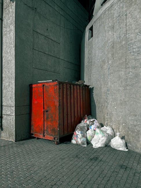 A large, rectangular red metal refuse skip with visible signs of rust and wear is positioned against a dark gray wall on a paved surface, near a light gray concrete wall. The skip is partially filled 