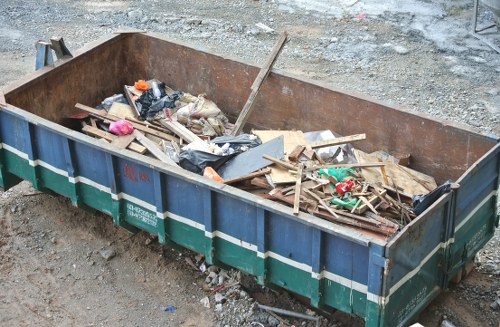 A large, rectangular metal skip with a rusted exterior, partially filled with mixed construction and household waste such as wooden planks, broken pieces of drywall, plastic bags, and miscellaneous debris. The skip is situated on a gravel surface, with a rough, uneven ground surrounding it, indicative of a waste clearance or building site in an urban area. The items inside the skip appear to be haphazardly piled, with some materials leaning over the sides. The background shows a patch of dirt or gravel, suggesting an outdoor setting typical for rubbish removal services in Belsizepark. This image reflects the type of waste that Belsizepark House Clearance manages through their comprehensive rubbish removal services, helping clients clear residential or commercial properties efficiently.
