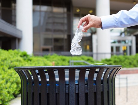 A person's hand, dressed in a light blue long-sleeve shirt, is seen dropping a crumpled, transparent plastic water bottle into a black, slatted outdoor rubbish bin situated on a paved area. The background features a modern building with large glass windows, some greenery, and a well-maintained courtyard in a residential or commercial area. The scene is well-lit with natural daylight, emphasizing the focus on waste disposal and clear separation of the bottle being correctly disposed of. This image illustrates proper rubbish disposal practices, relevant to services offered by Belsizepark House Clearance for waste and rubbish removal in the Belsizepark area, within the context of sustainable waste management.